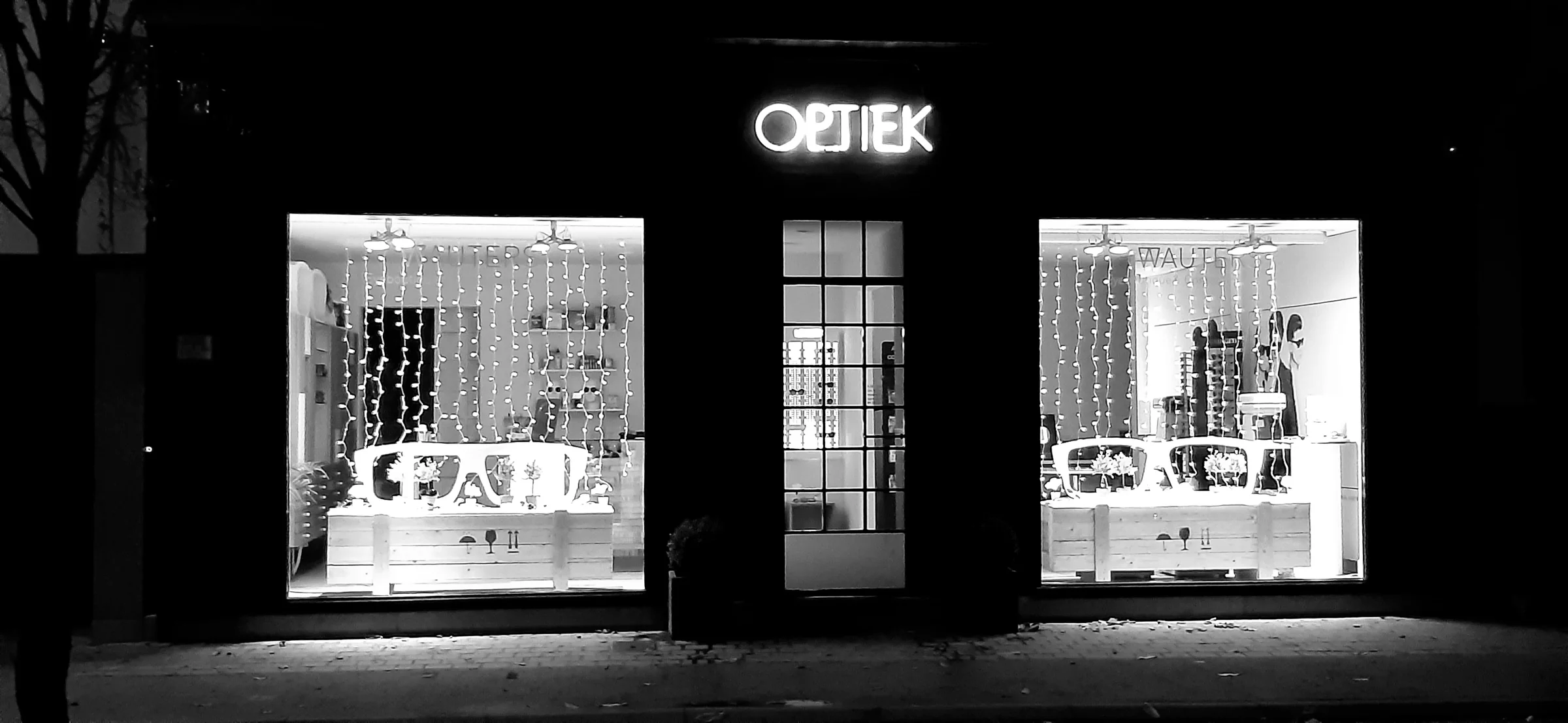 De gevel van Optiek Wauters bij avond: het verlichte OPTIEK-uithangbord boven de deur en twee etalages vol monturen